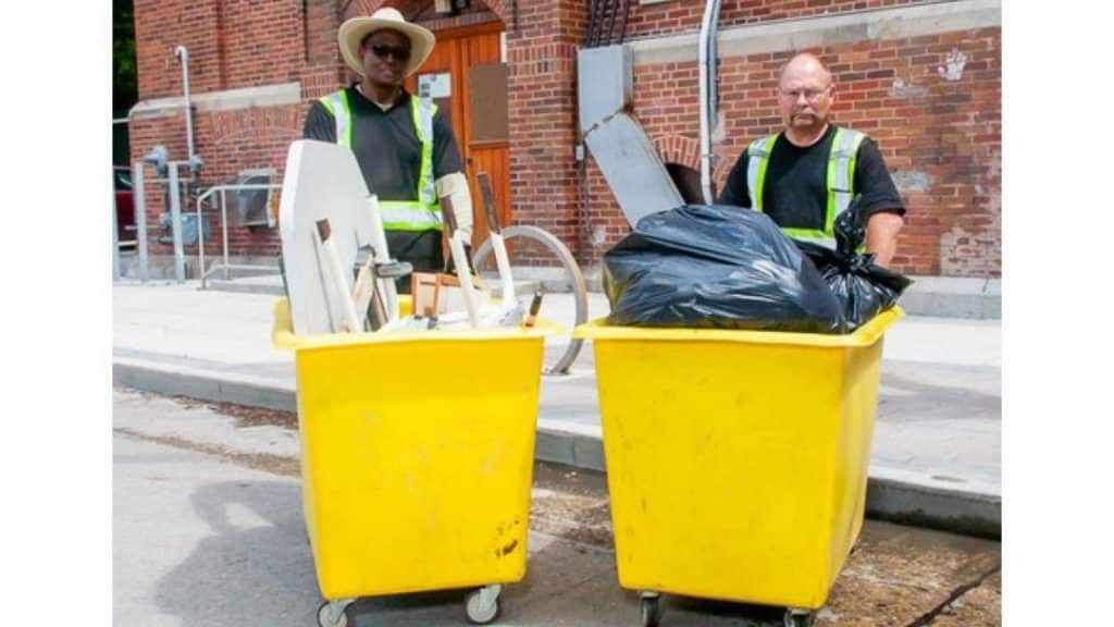 Two waste management workers standing outdoors with large yellow bins filled with garbage and debris in front of a brick building.